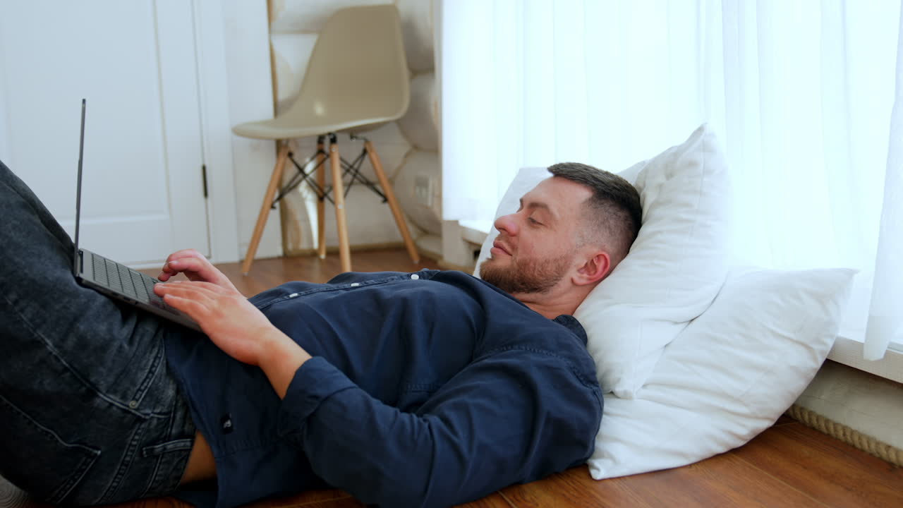 Relaxed Caucasian male lies on the floor with two pillows under his head. Man holds a laptop on his belly and touches keyboard.