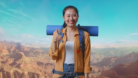 asian female hiker with mountaineering backpack smiling and showing thumbs up gesture to camera while traveling at the top of mountain