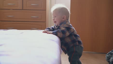 fair haired baby leans on soft bed with pink plaid in room