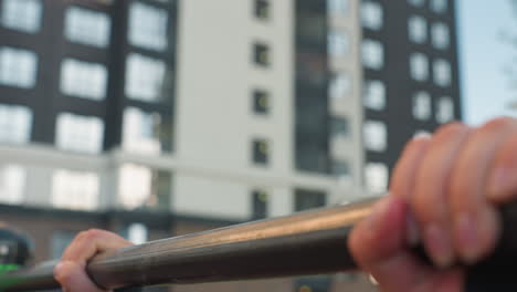 close up hands of fit coach gripping horizontal pull up bar in urban park during intense exhale with chest contact ensuring balanced form and controlled muscle engagement against skyscraper backdrop