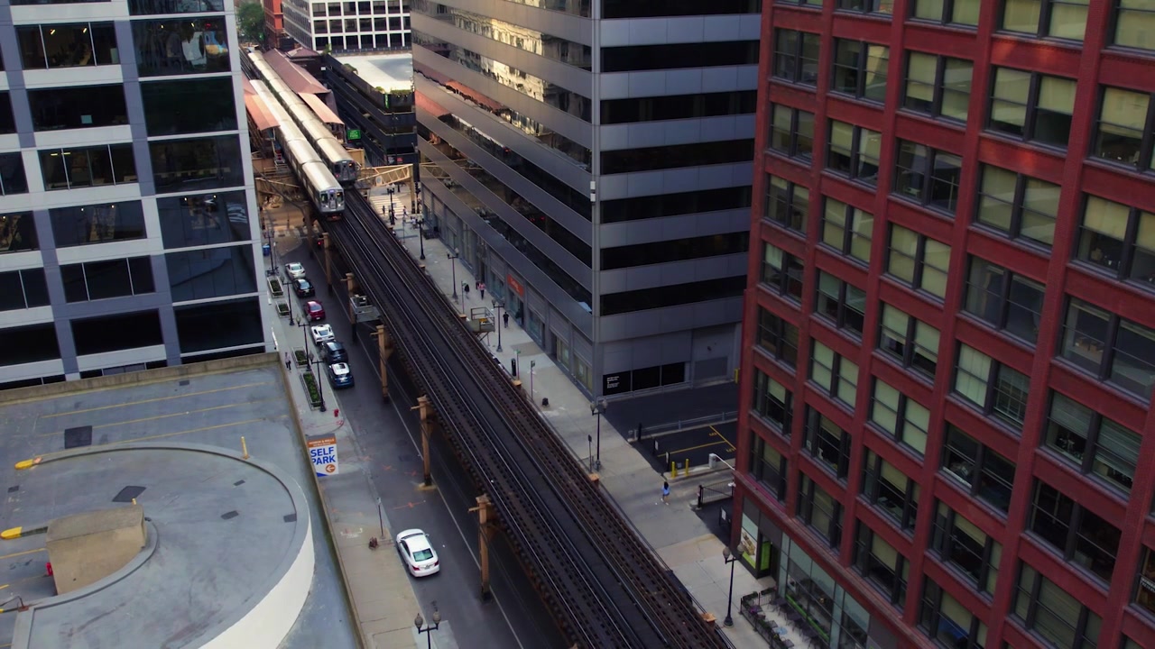 Tomada de drone acima de um trem da CTA viajando ao lado de uma estação elevada pelo centro de Chicago, cercado por edifícios de escritórios, ruas e arquitetura urbana densa