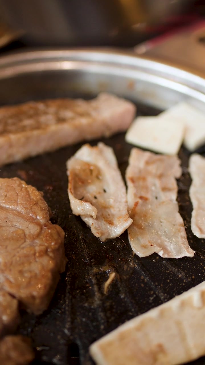 Una mano gira fette di maiale con le pinze su una calda griglia coreana per barbecue, illuminazione calda, profondità di campo ridotta.