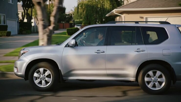 SUV argenté circulant près de la caméra dans une rue de banlieue, un homme en chemise sombre au volant.