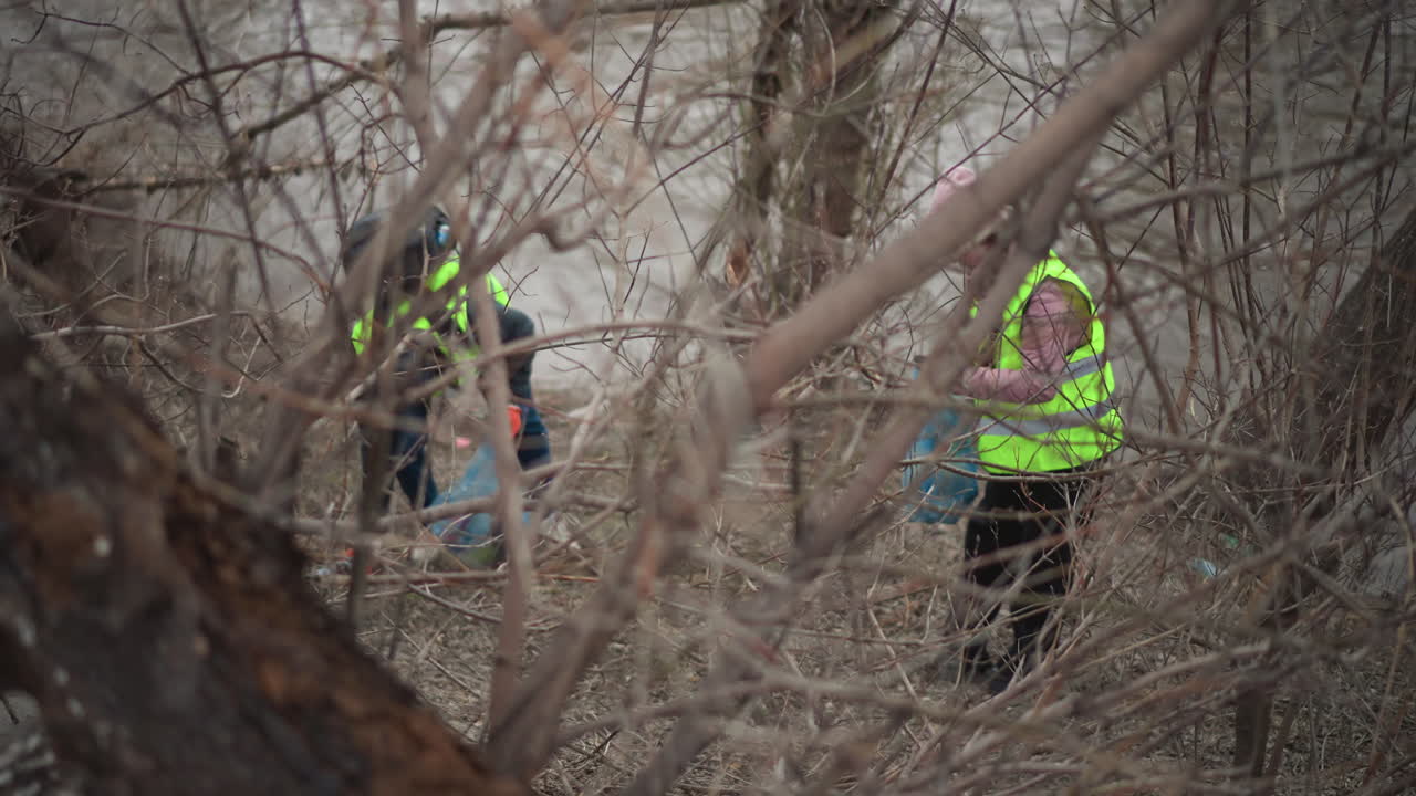 Volunteers in bright reflective vests and gloves picking up plastic bottles and trash along riverbank through leafless branches during environmental cleanup effort on cold overcast day to protect nature