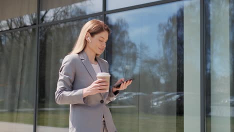 hermosa mujer de negocios de mediana edad mensajería en el teléfono celular sosteniendo el café de la mañana mientras camina por la calle de la ciudad. mujer mirando hacia adelante y lejos pensativo. redes sociales. teléfonos inteligentes. aplicaciones.