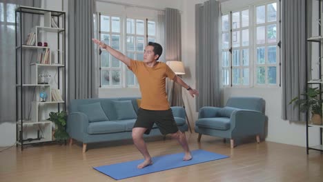 hombre asiático haciendo yoga en warrior ii en la alfombra en casa