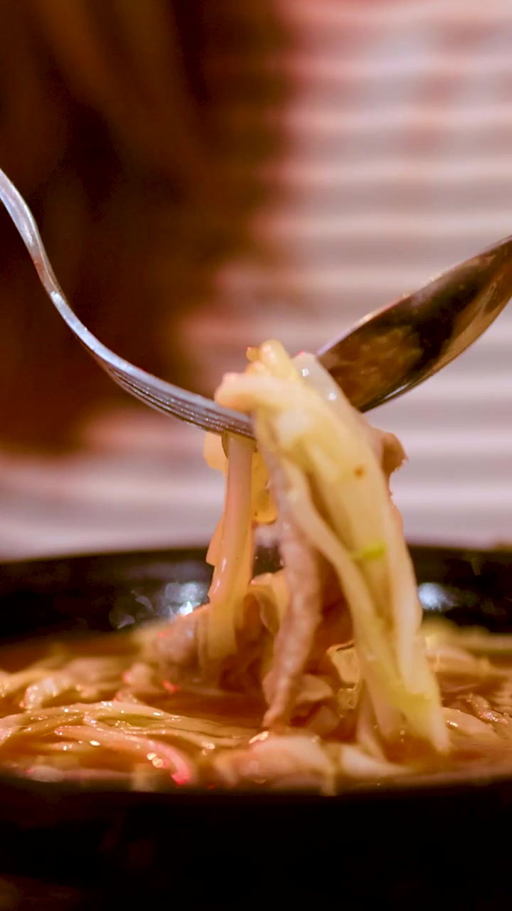 Primo piano di una forchetta e un cucchiaio che sollevano noodles e carne da una ciotola fumante di pho vietnamita.