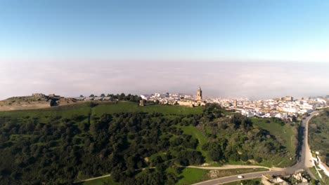 Aerial-dolly-shot-over-the-landscape-of-medina-sidonia-in-cadiz-spain-with-a-view-of-the-busy-country-road-along-the-hills-with-trees-and-the-historic-old-town-with-white-buildings-on-a-sunny-day