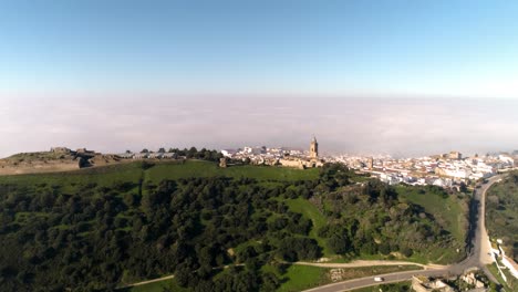 tiro aéreo sobre el paisaje de medina sidonia en cádiz españa con vistas a la concurrida carretera rural a lo largo de las colinas con árboles y al casco histórico con edificios blancos en un día soleado
