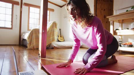 mujer de raza mixta relajada practicando yoga, arrodillándose y estirándose en un dormitorio de cabaña soleado