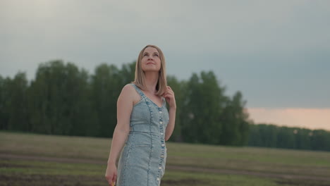 woman adjusts hair while looking into distance over open grassy field under cloudy summer sky wearing denim dress holding pinwheel capturing contemplative mood and gentle breeze
