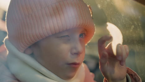 close-up of a young girl wearing a pink knit hat and scarf, tapping on a car window with her finger, one eye closed in a moment of playful curiosity