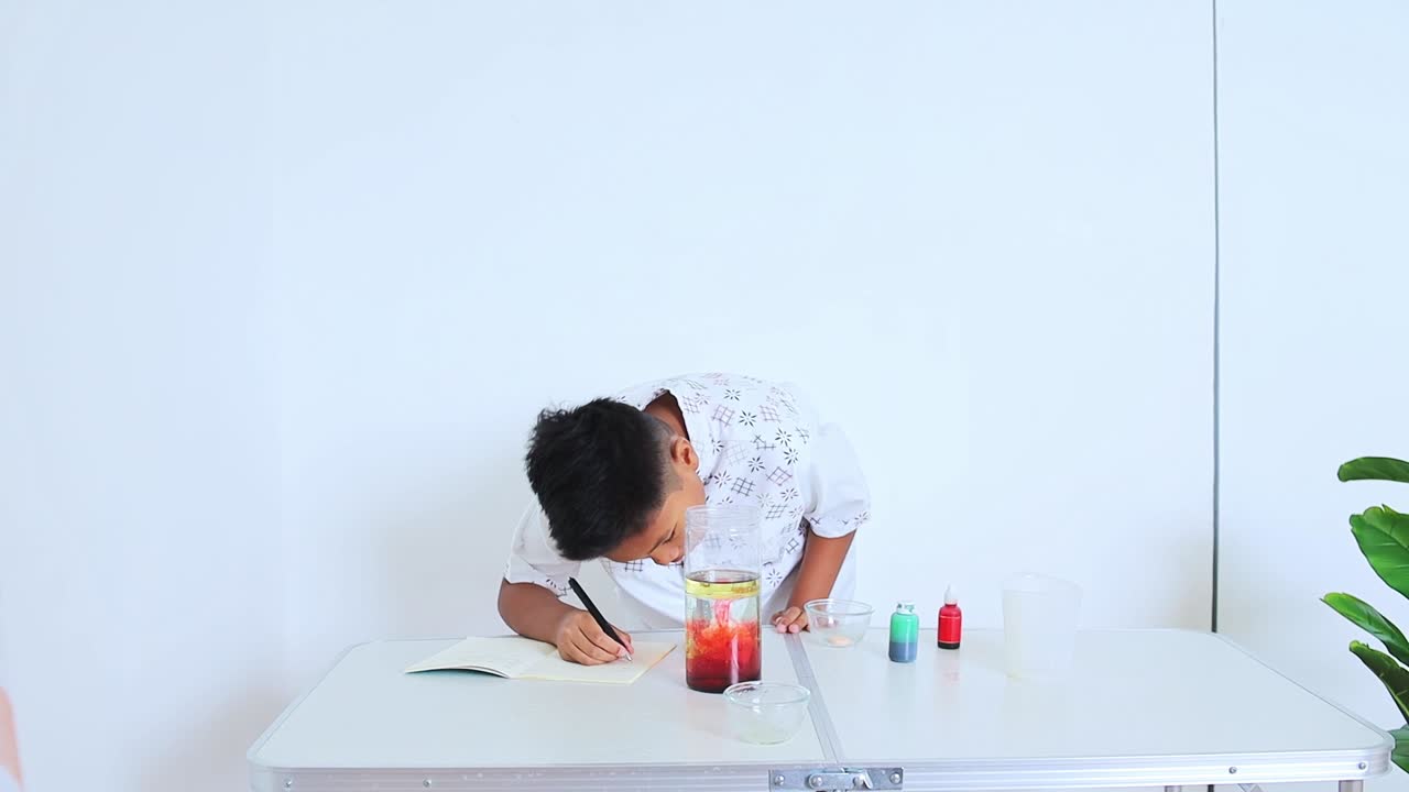 Ragazzo asiatico che osserva un esperimento con lampada lava colorata e scrive i risultati nel quaderno a casa