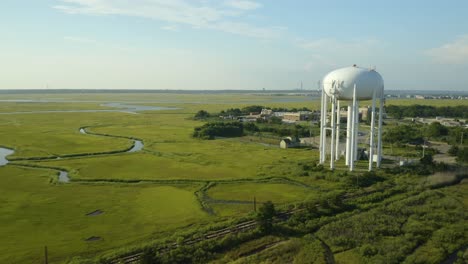 Aerial,-tall-white-water-tank-tower-in-green-swampland,-sunny-day