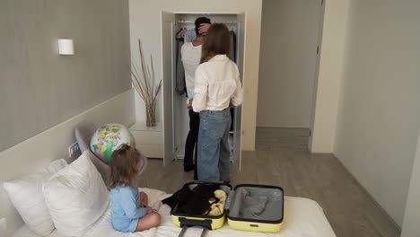 Young-modern-caucasian-couple-gets-ready-for-trip-together-with-their-little-daughter,-packs-suitcase-having-fun,-smiles,-laughs