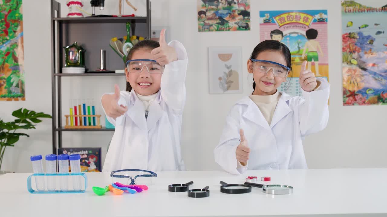 Kaksi nuorta aasialaista tyttöä, yllään laboratoriotakit ja suojalasit, hymyilevät ja näyttävät peukkuja ylöspäin.