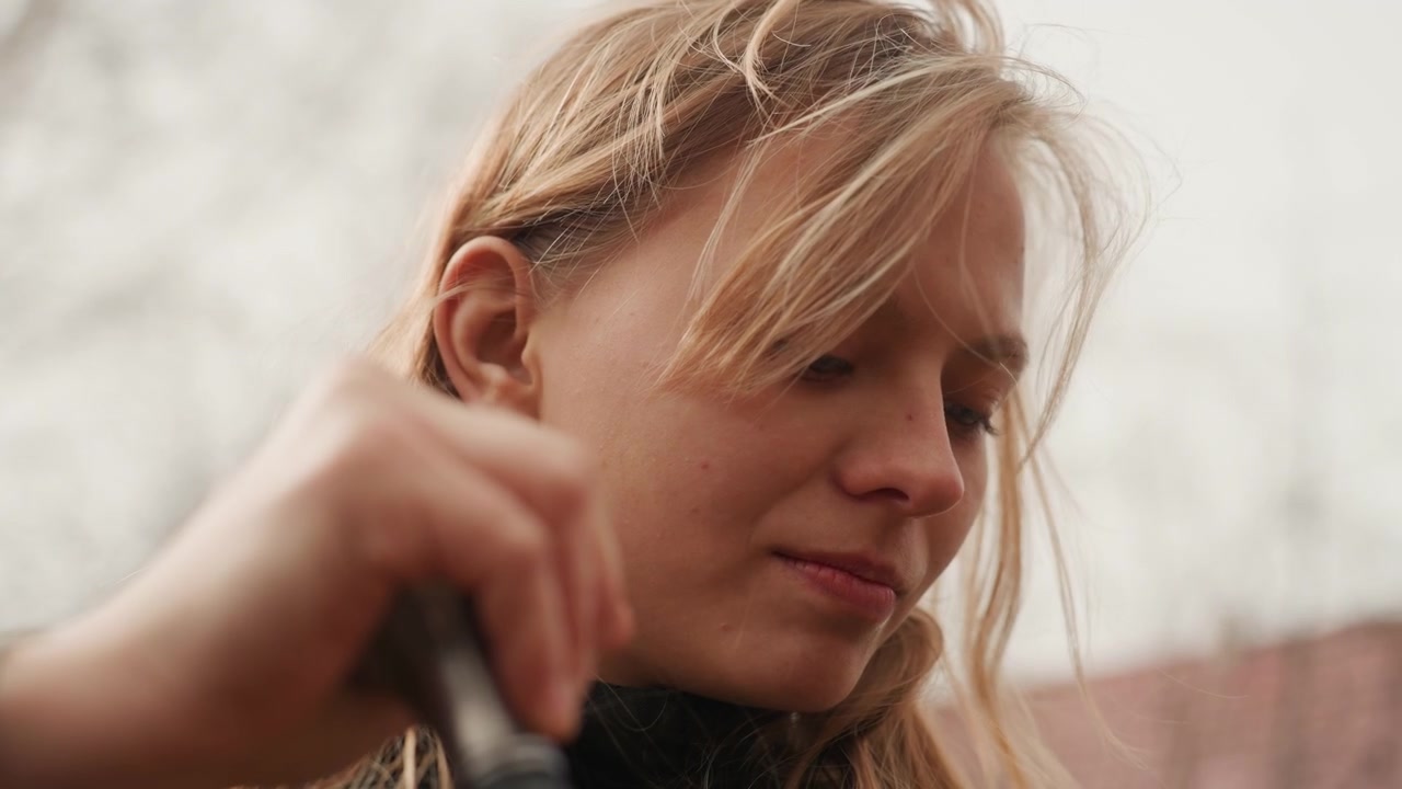 Young woman in overcast garden grips outdoor tool handle with both hands while looking down, damp hair falling across forehead, calm focused expression, soft natural light, quiet seasonal work