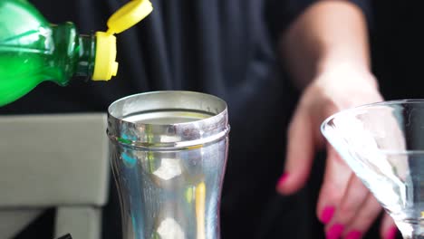 home bartender pours lemon or lime juice from green squeeze bottle in a silver cobbler shaker to make a cocktail in a martini glass, closeup of woman’s hands with pink nail polish