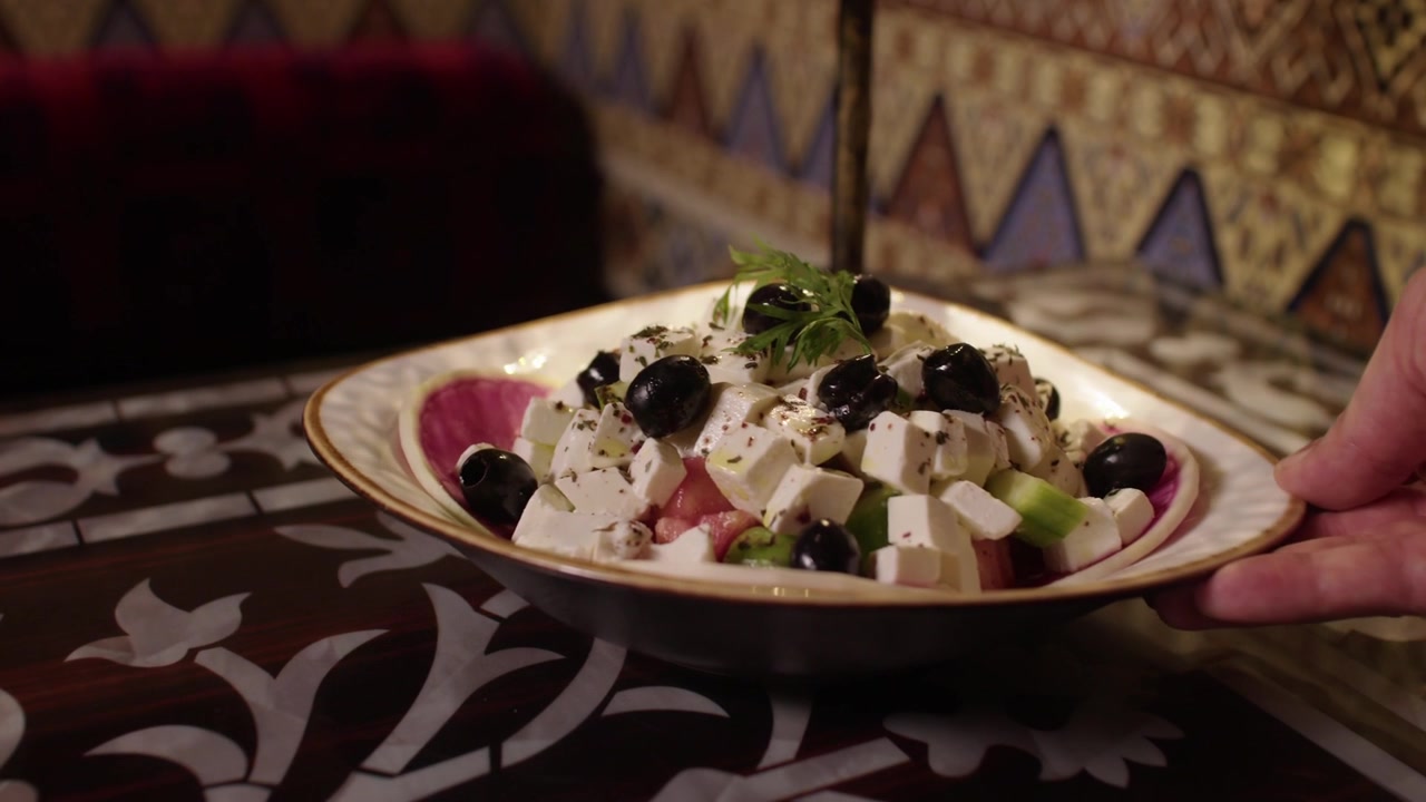 Una mano posa un'insalata fresca con feta e olive su un tavolo da ristorante decorato.
