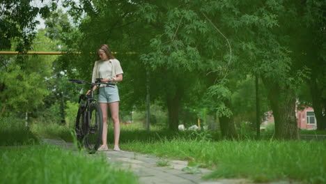 woman holding black gloves approaches her parked bicycle on paved path, wears her gloves, background features lush greenery, people passing by in distance, and gentle breeze moving leaves around