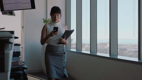 mujer de negocios asiática que usa un teléfono inteligente caminando por la oficina enviando mensajes de texto una mujer ejecutiva exitosa chequeando correos electrónicos en el teléfono móvil en el lugar de trabajo