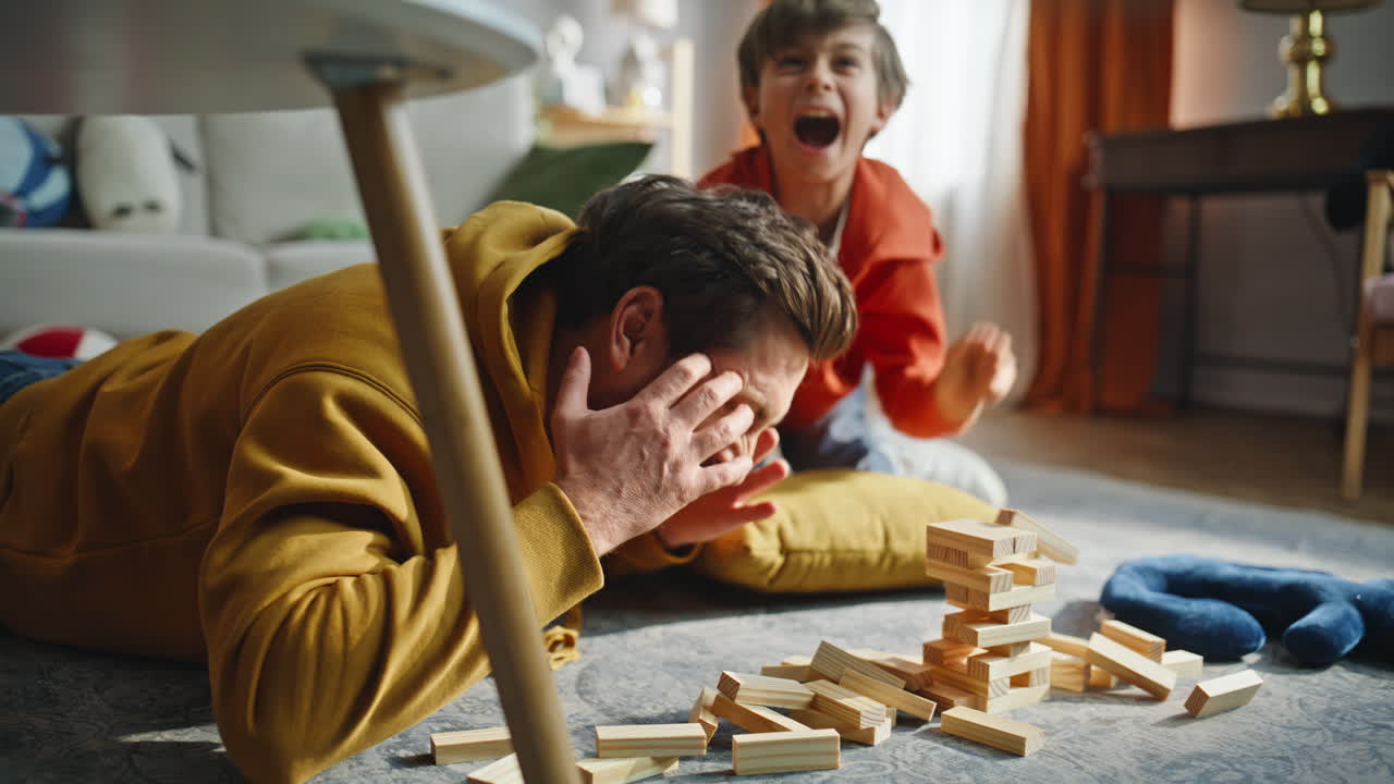 Father ruined jenga tower playing with happy little boy lying home floor closeup
