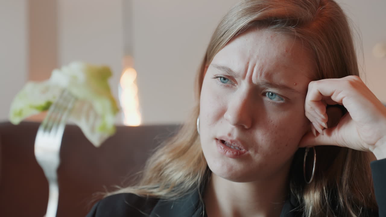 White woman frowning at lettuce fork, reluctant diner evaluating salad during urban caf break, warm lamp light, closeup facial expression showing doubt, subtle tension and hesitation before bite