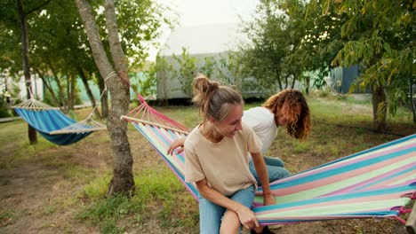 un tipo con gafas, con el cabello rizado en una camiseta blanca se acerca a la hamaca con su novia y se acuestan en ella juntos. un tipo golpea a su novia en la cabeza. descanso en la casa de campo