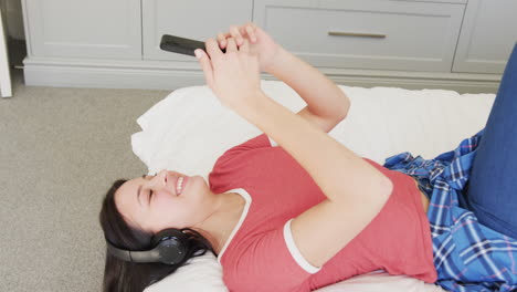 happy biracial woman in headphones lying on bed using smartphone, slow motion