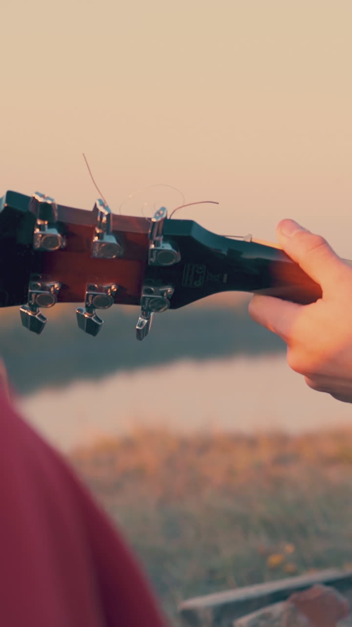 man plays guitar among tourists in camp on steep river bank in autumn evening at sunset close backside view