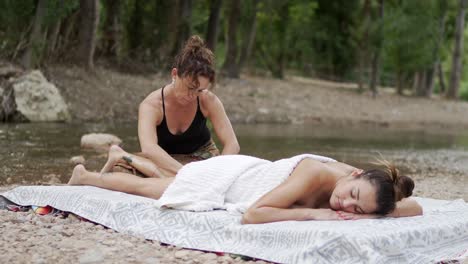 masseuse rubbing feet of woman lying near rippling water of pond