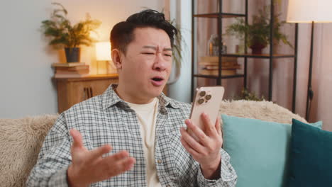 upset displeased chinese man using smartphone reading bad news sitting on sofa couch in living room