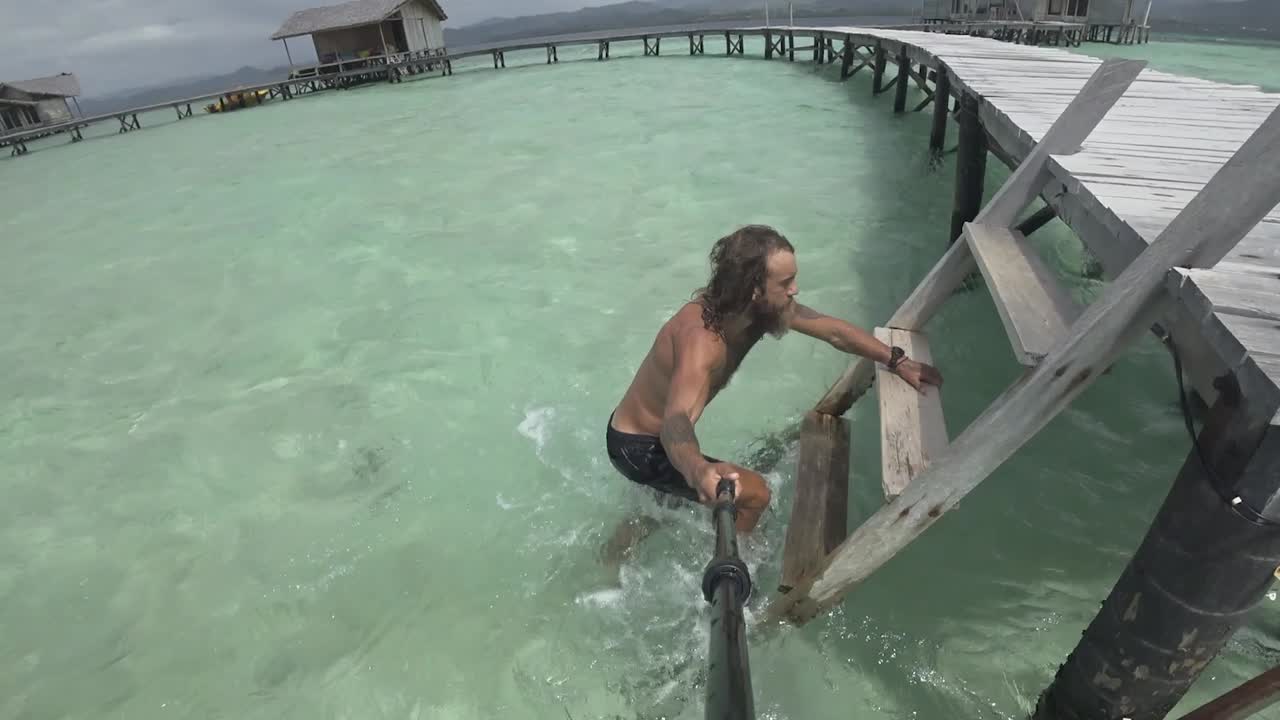 A young, fit and strong man is walking in the shallow water, walks up the small ladder onto the wooden jetty and starts running with the paradise island of white sand in the background