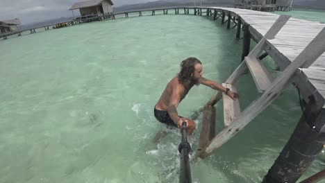 a young, fit and strong man is walking in the shallow water, walks up the small ladder onto the wooden jetty and starts running with the paradise island of white sand in the background