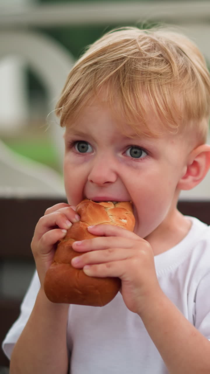 A young boy is seen seated and focused as he tries to take a larger bite of a sausage roll, there's a partial view of someone nearby in a white outfit