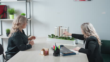 saleswoman gesturing with pencil while explaining modern residential building miniature to customer at office desk, with focus on architectural model
