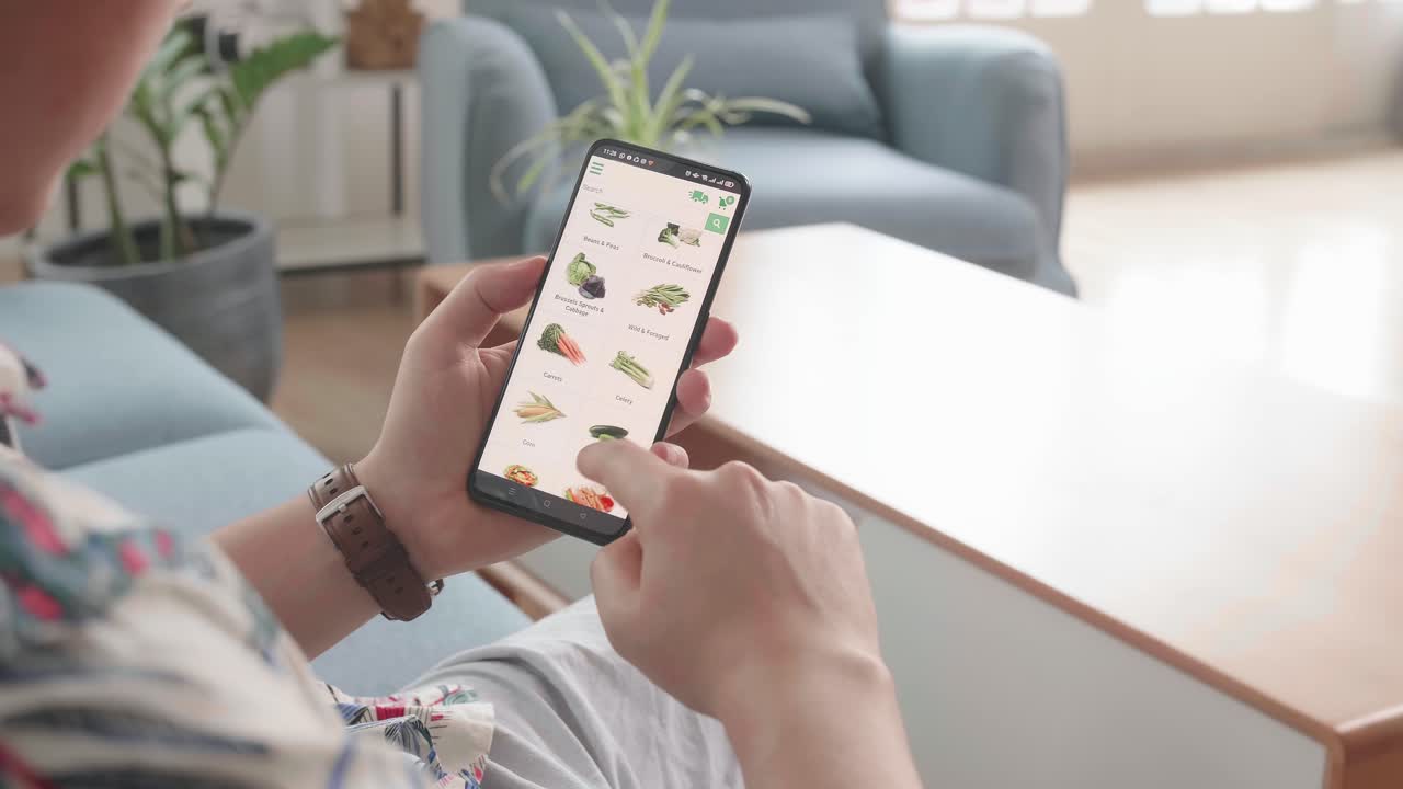 Man Sitting On Couch Using Smartphone Buys In Internet Shop. Male Selects Vegetables In Grocery Online Store