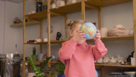 blonde little girl playing with globe sitting at a table where are drawings of the earth in a craft workshop