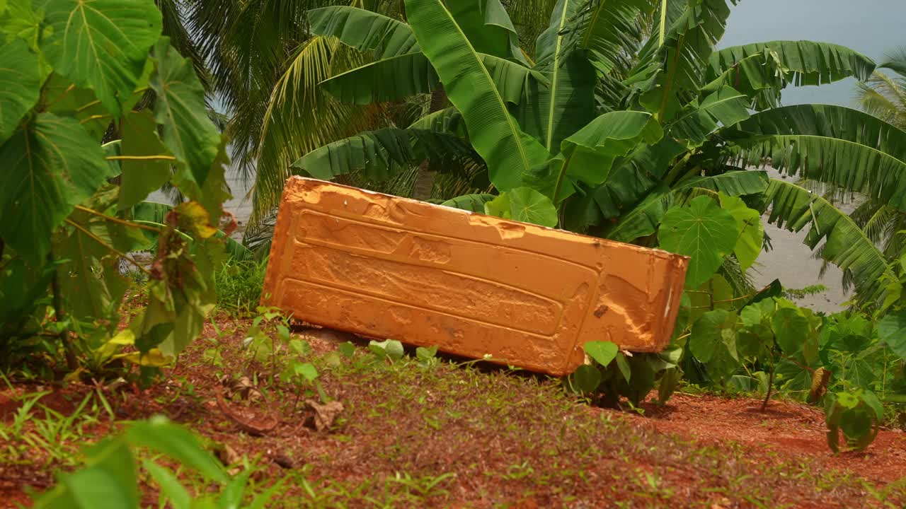 A still shot of an orange styrofoam block lies on red earth amid tropical growth, its bright surface clashing with nature’s palette near the Mauban Port, Quezon Province Philippines