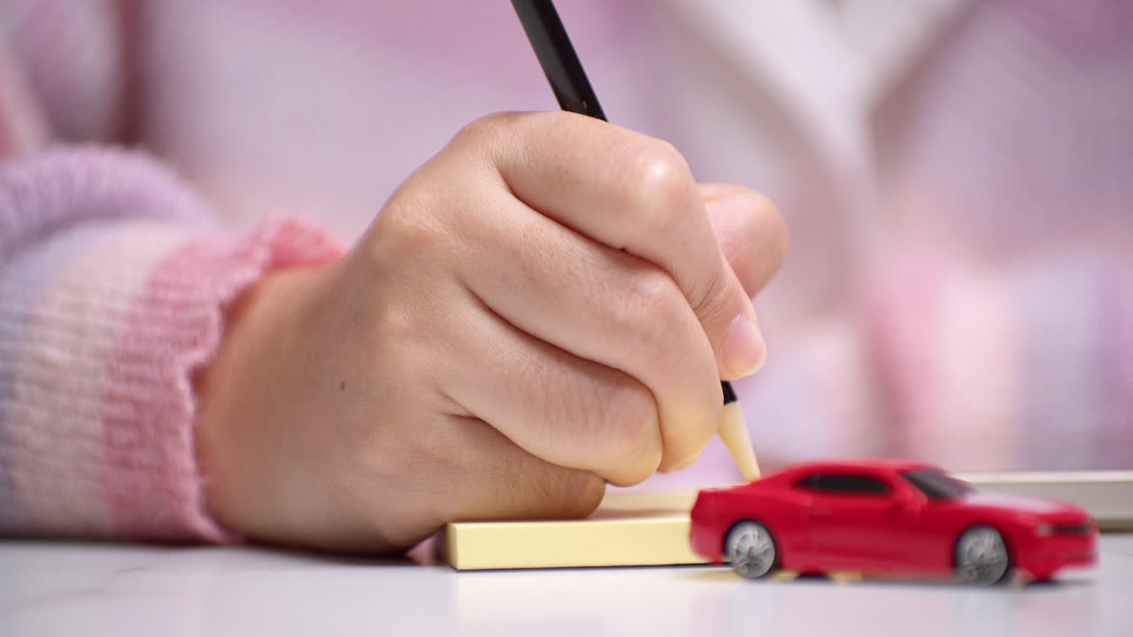 primer plano de una mano sosteniendo un lápiz y escribiendo algo en un papel amarillo, detrás de un coche de juguete rojo que significa financiación de préstamo de coche