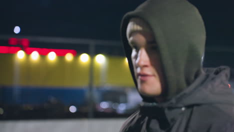 man in hoodie with focused expression during nighttime activity on outdoor urban field, background features illuminated fence, vibrant yellow and red lights