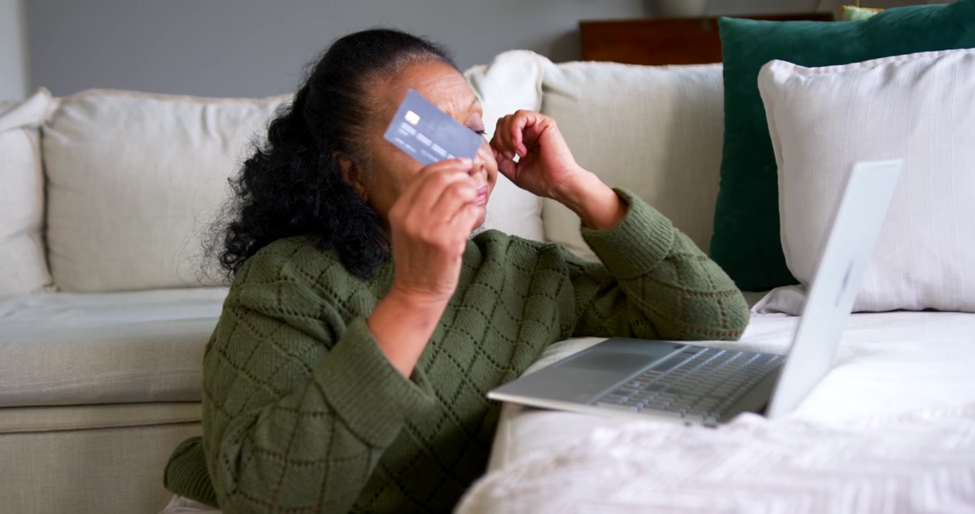 Senior African American woman in green knit pausing typing at laptop on couch, picking card, paying