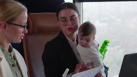 Meeting-of-two-colleagues-Business-Woman-brunette-girl-in-round-glasses-and-a-business-suit-holds-her-small-infant-child-while-communicating-on-serious-issues-with-her-colleagues-at-the-office-table