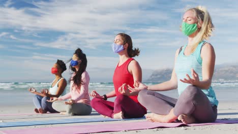 group of diverse female friends wearing face masks meditating at the beach