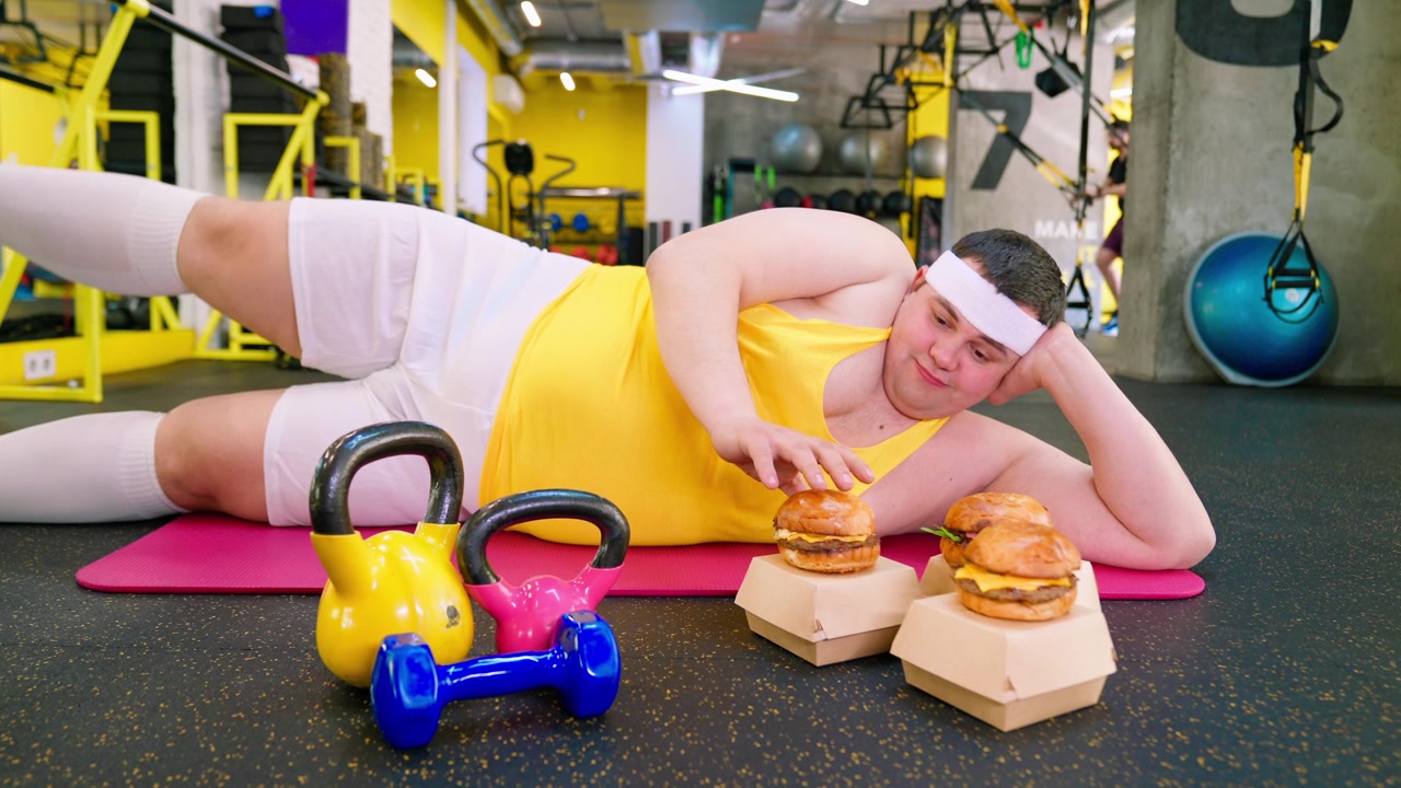 Uomo pigro e in sovrappeso sdraiato sul tappetino, tentato dagli hamburger in palestra