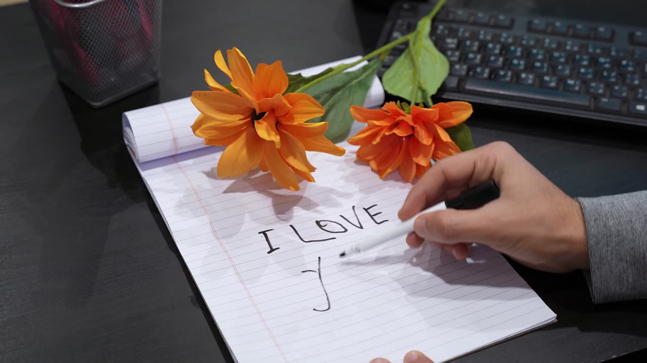 Close-up van een hand die ‘I LOVE YOU’ schrijft en een hart tekent op gelinieerd notitiepapier, oranje bloemen naast een laptoptoetsenbord op een donker bureau, romantisch Valentijnsconcept met een kopieerruimte