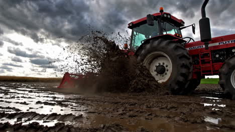 red tractor navigating a muddy field under a cloudy sky