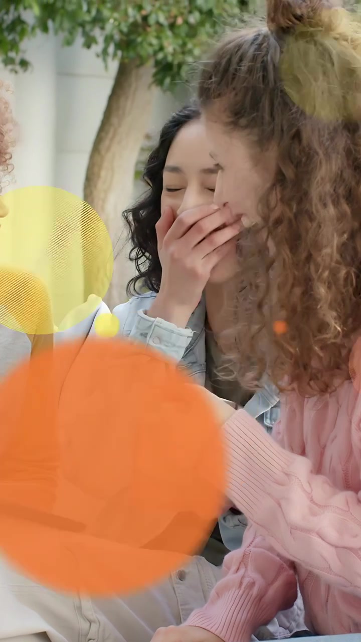 Vertical video: Friends laughing over tablet, center woman holding mug covering mouth, social bokeh