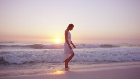 Hermosa-Mujer-Con-Vestido-Blanco-Caminando-A-Lo-Largo-De-La-Costa-En-La-Playa-Al-Atardecer-En-Cámara-Lenta-Dragón-Rojo