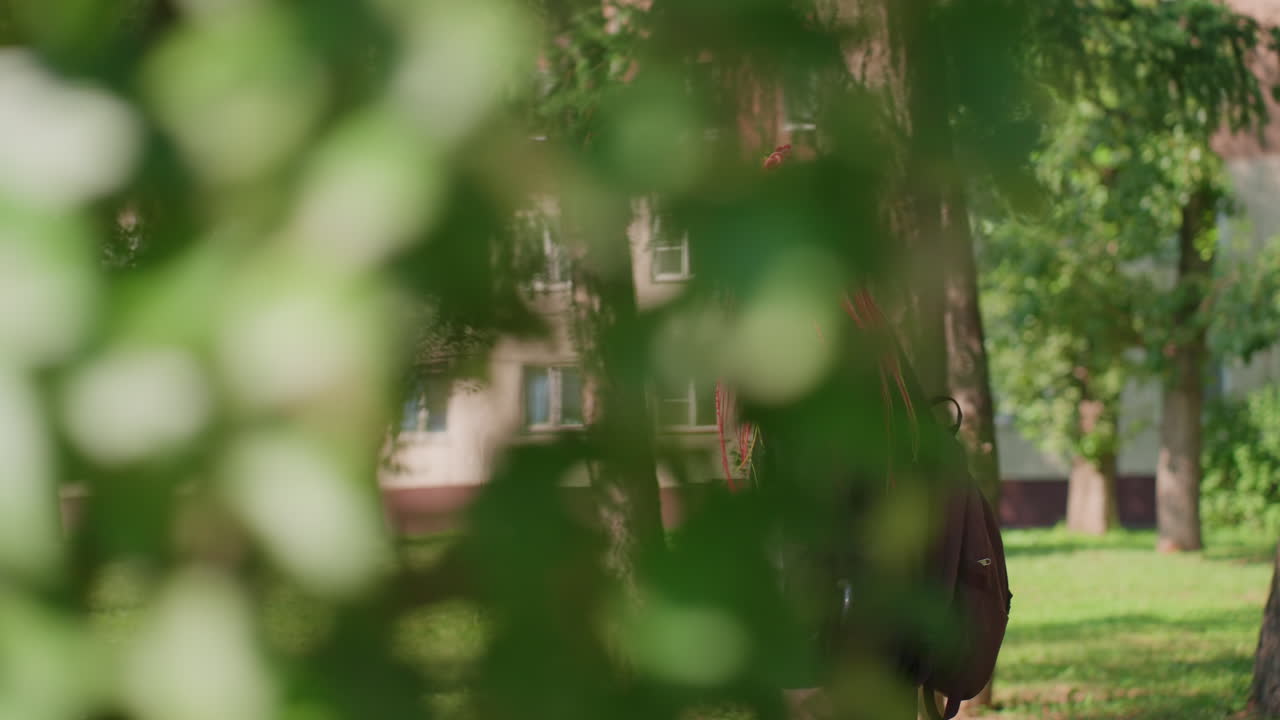 보케가 도시의 자연 환경 장면으로 희미하게 사라지고, 부드러운 초점이 나무와 건물 근처를 걷는 여성으로 이동하며, 풍성한 나뭇잎와 도시 건축물 사이에서 네온 브레이드 머리를 한 여성이 편안하게 걷는 장면입니다.